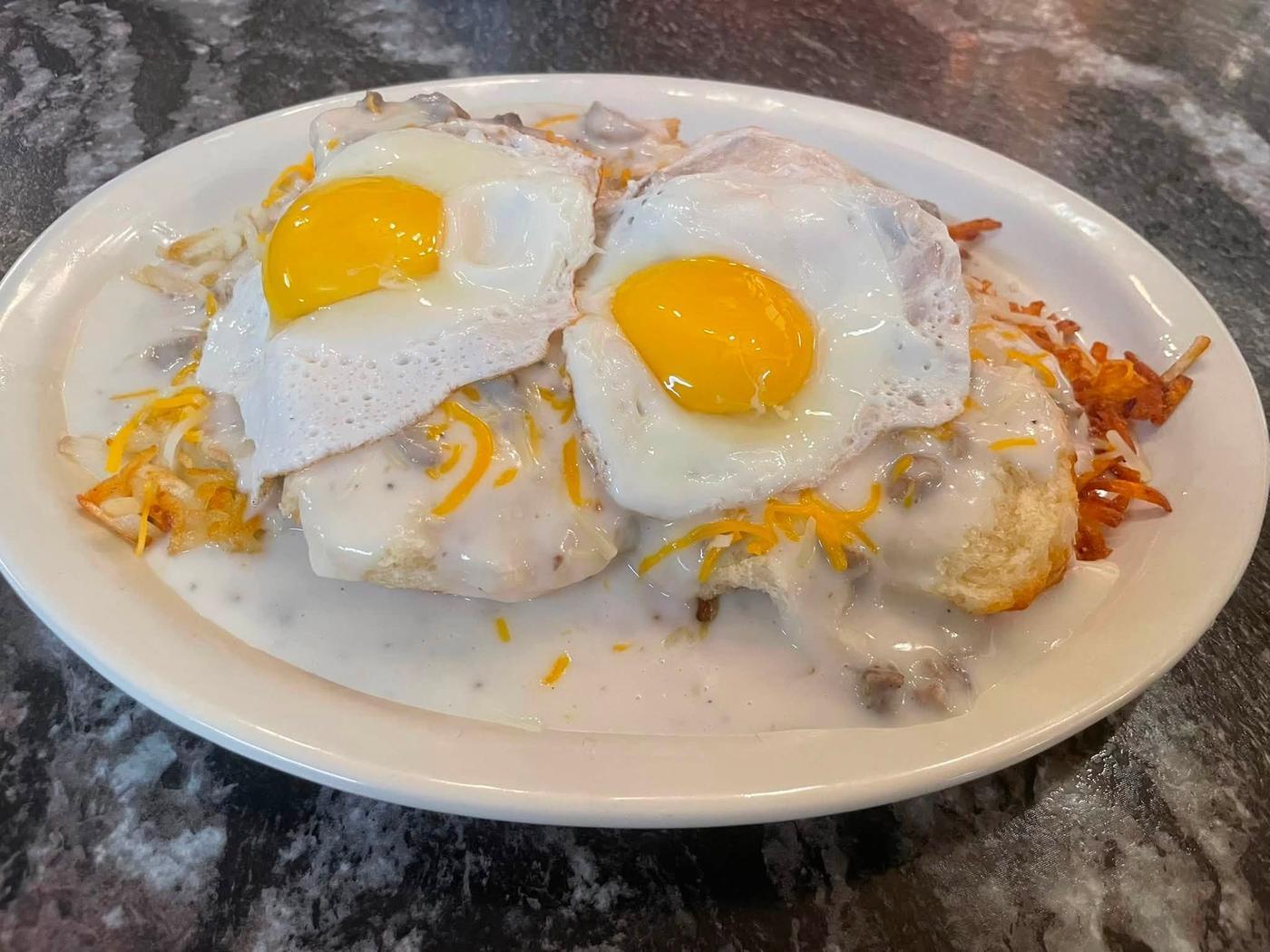 Two biscuits under country gravy with two fried eggs, shredded cheese and a pile of hash browns.