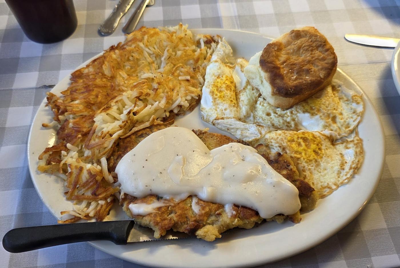 A chicken fried steak under white country gravy with hash browns, three eggs and a tall biscuit, on a gingham tablecloth.
