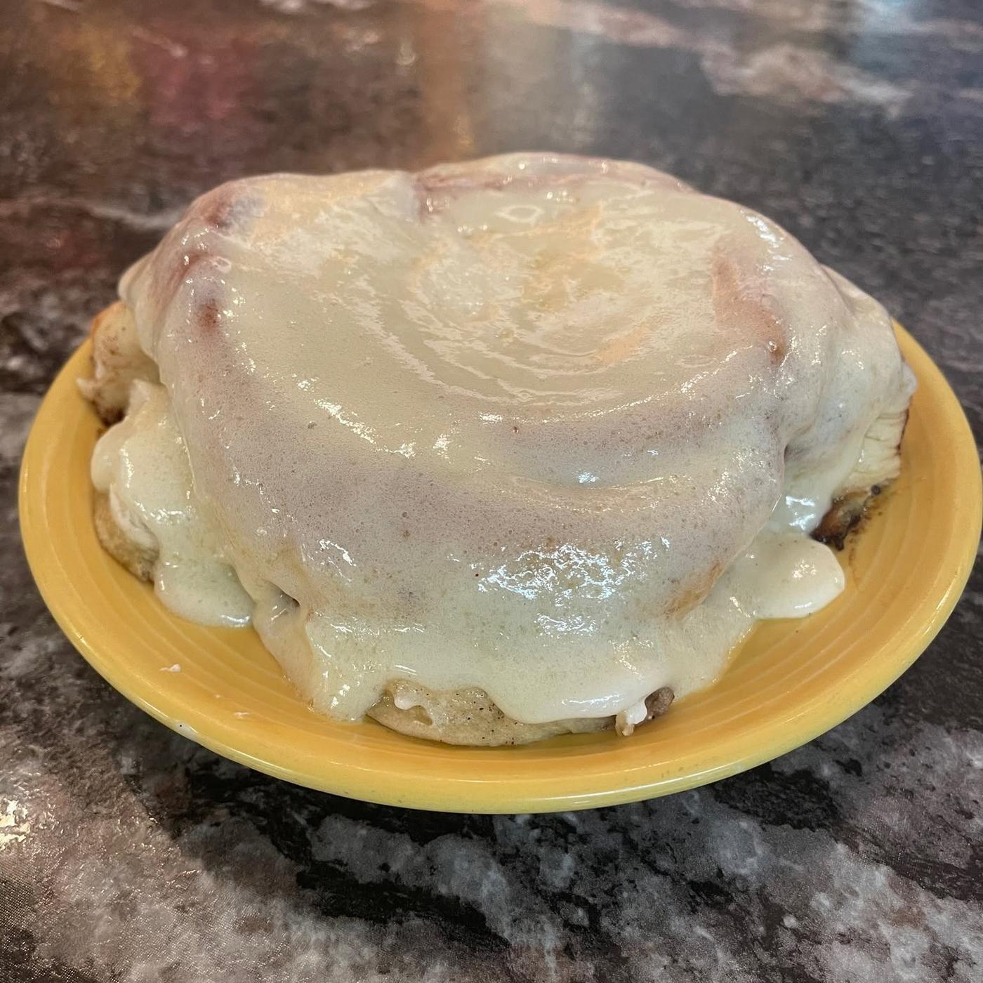 A cinnamon roll wider than the plate it sits on, under cream cheese icing running down the sides.