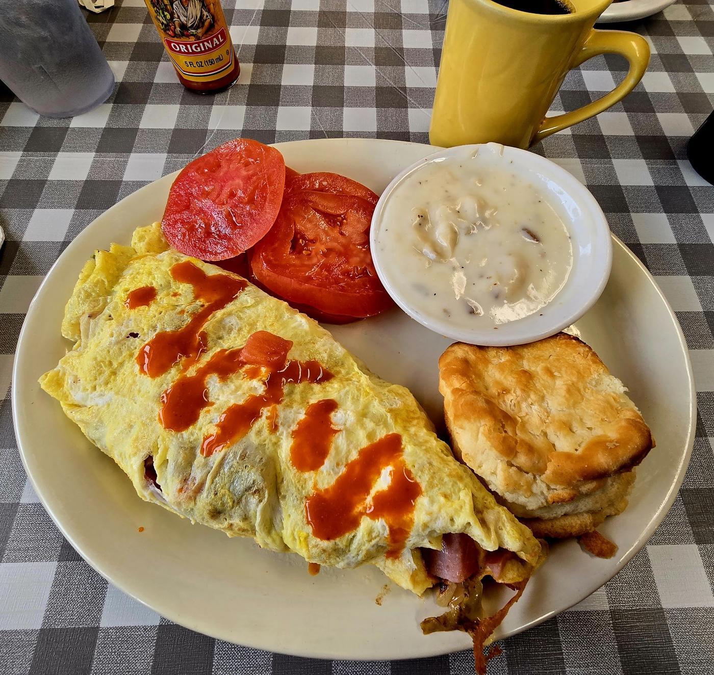 A folded three egg omelette with hot sauce across it, sliced tomato, a ramekin of sausage gravy and a biscuit.